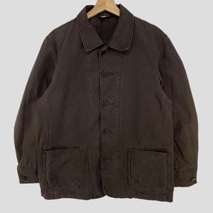 May include: A brown jacket with a button-up front, two large front pockets, and a collar. The jacket is hanging on a wooden hanger against a light gray background.