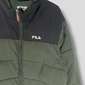 May include: A green and black FILA puffer jacket. The jacket has a high collar and a full-length zipper. The FILA logo is displayed in white on the chest. The jacket is hanging on a black hanger.