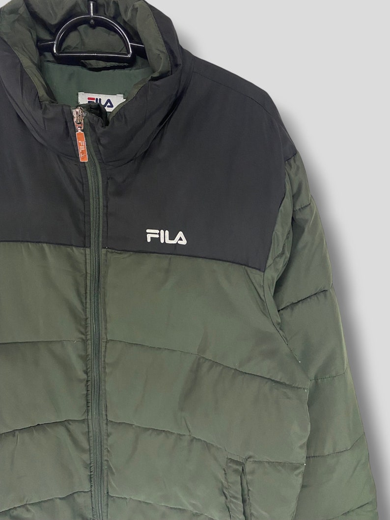 May include: A green and black FILA puffer jacket. The jacket has a high collar and a full-length zipper. The FILA logo is displayed in white on the chest. The jacket is hanging on a black hanger.