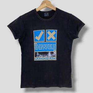 May include: Black t-shirt with a graphic featuring a blue tick and a cross, accompanied by the text "MOST PEOPLE GAZE NEITHER INTO THE PAST NOR THE FUTURE: THEY EXPLORE NEITHER TRUTH NOR LIES. THEY GAZE AT THE TELEVISION." and the band name "RADIOHEAD."