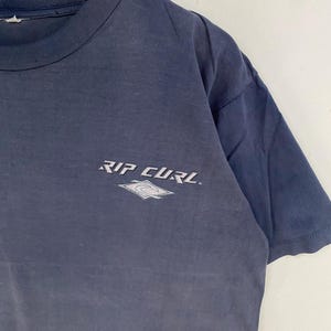 May include: A navy blue t-shirt with the text "RIP CURL" in white, along with a white and gray logo. The shirt is made of a soft material and has short sleeves. The shirt is hanging on a wooden hanger.