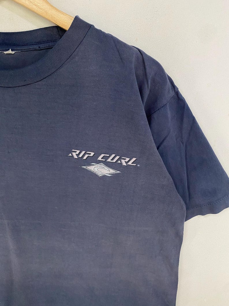 May include: A navy blue t-shirt with the text "RIP CURL" in white, along with a white and gray logo. The shirt is made of a soft material and has short sleeves. The shirt is hanging on a wooden hanger.