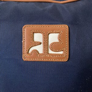May include: A navy blue bag with a brown leather patch. The patch has a white interior with a brown border and stitched detailing. The bag has a brown trim along the top edge.