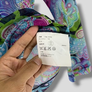 Puede incluir: Primer plano de una prenda con estampado de cachemir en colores vibrantes. La tela presenta una mezcla de tonos azules, verdes, morados y rosas. Se ve una etiqueta con texto japon&eacute;s, incluido el c&oacute;digo de producto TM-T009 y la composici&oacute;n del material, 100% poli&eacute;ster.