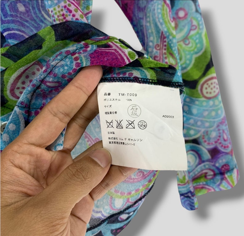 Puede incluir: Primer plano de una prenda con estampado de cachemir en colores vibrantes. La tela presenta una mezcla de tonos azules, verdes, morados y rosas. Se ve una etiqueta con texto japon&eacute;s, incluido el c&oacute;digo de producto TM-T009 y la composici&oacute;n del material, 100% poli&eacute;ster.