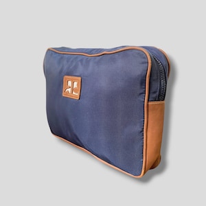 May include: Navy blue travel bag with brown leather accents. The bag features a brown leather patch with a logo and a brown zipper. The bag is rectangular and has a rounded top.