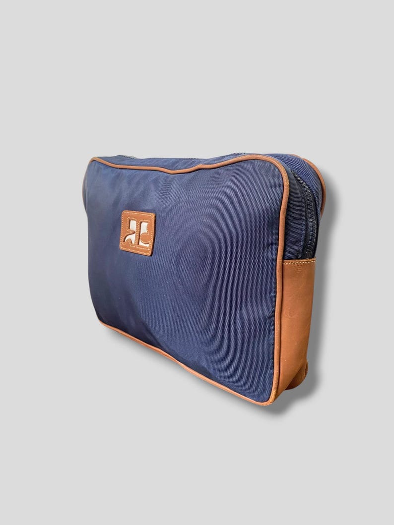 May include: Navy blue travel bag with brown leather accents. The bag features a brown leather patch with a logo and a brown zipper. The bag is rectangular and has a rounded top.