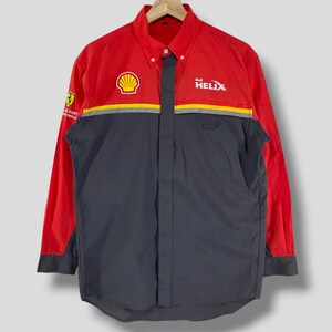Puede incluir: Una camisa de manga larga con botones de color rojo y negro con el logotipo de Shell y el texto "Shell Helix" en la parte delantera. La camisa también tiene un logotipo de Ferrari en la manga izquierda.