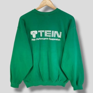 May include: A bright green crewneck sweatshirt featuring the white text TEIN and the words High Performance Suspension printed on the chest. The garment has long sleeves with ribbed cuffs and a ribbed neckline, displayed on a wooden hanger.