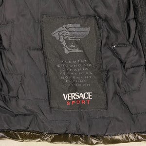 May include: A black quilted jacket with a black label that says "VERSACE SPORT" and has the text "ELEMENT ERGONOMIC DYNAMIC TECHNICAL MOVEMENT FUTURE OUTDOOR" in white.