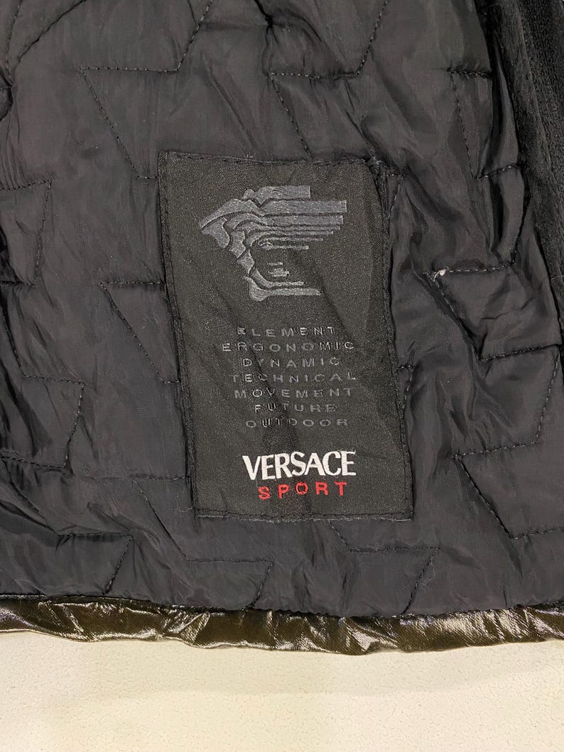 May include: A black quilted jacket with a black label that says "VERSACE SPORT" and has the text "ELEMENT ERGONOMIC DYNAMIC TECHNICAL MOVEMENT FUTURE OUTDOOR" in white.
