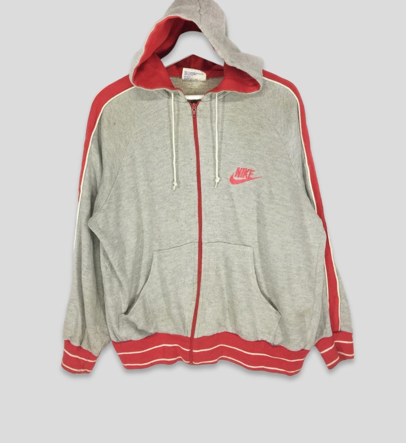 K&ouml;nnte beinhalten: Ein grauer Kapuzenpullover mit Rei&szlig;verschluss, roter Innenseite, roten Streifen an den &Auml;rmeln und roten Verzierungen an den B&uuml;ndchen und am Bund. Das Nike-Logo ist rot auf die Brust gestickt. Der Hoodie hat eine Kapuze mit Kordelzug und eine Vordertasche.