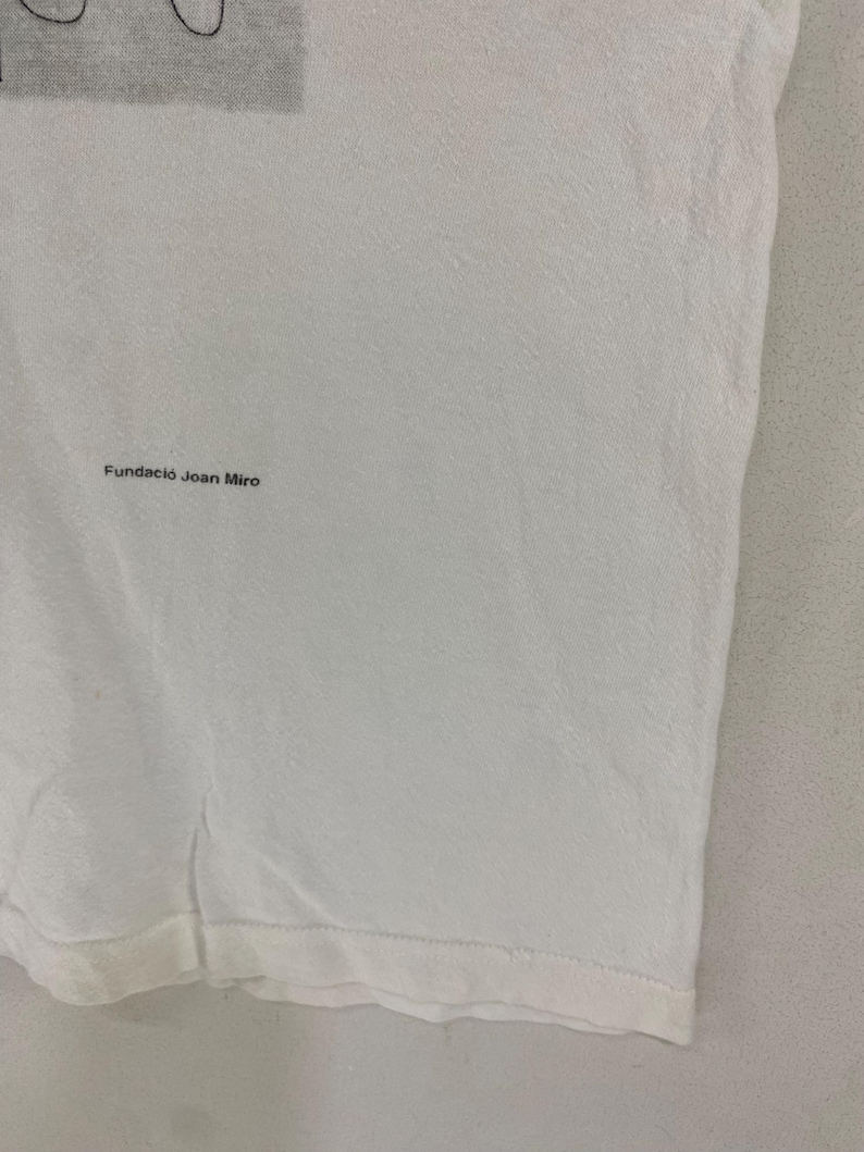 Puede incluir: Una camiseta blanca con el texto "Fundaci&oacute; Joan Mir&oacute;" impreso en negro. La tela tiene una textura sutil. El dobladillo inferior de la camiseta es visible.