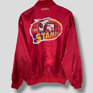 Könnte beinhalten: Rote Vintage-Jacke mit Reißverschluss und geripptem Saum und Bündchen. Auf der Rückseite der Jacke befindet sich eine farbenfrohe Grafik mit dem Sony-Logo, dem Wort "STAMINA" und dem Text "TRY".