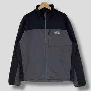 May include: A two-tone jacket from The North Face, in black and gray. The jacket has a full-length zipper, a small chest pocket, and the brand's logo on the chest. The jacket is hanging on a wooden hanger.