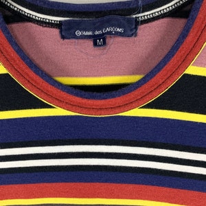 May include: Close-up of a striped shirt with a round neckline. The shirt features horizontal stripes in various colors, including black, yellow, blue, red, and white. The label reads "Comme des Garcons" and size "M".