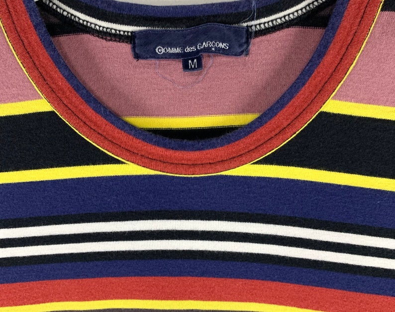 May include: Close-up of a striped shirt with a round neckline. The shirt features horizontal stripes in various colors, including black, yellow, blue, red, and white. The label reads "Comme des Garcons" and size "M".
