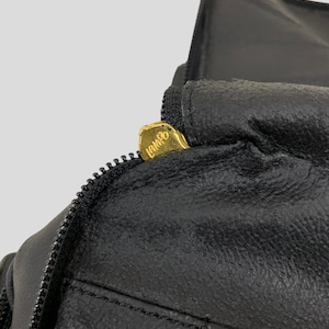 May include: Close-up of a black leather item with a zipper. The zipper pull is gold-colored and has the word "LANCO" engraved on it. The leather appears to be textured and stitched.
