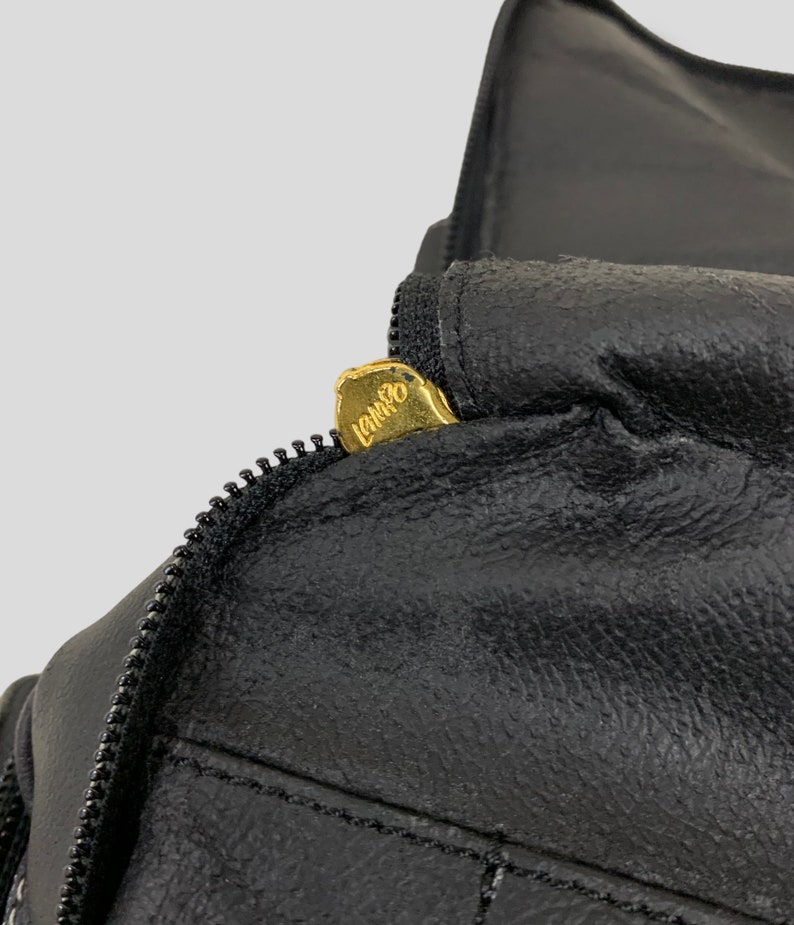 May include: Close-up of a black leather item with a zipper. The zipper pull is gold-colored and has the word "LANCO" engraved on it. The leather appears to be textured and stitched.