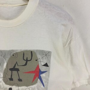 Puede incluir: Camiseta blanca con un dise&ntilde;o abstracto. El dise&ntilde;o incluye un fondo gris, formas negras, una estrella roja y una estrella azul. La camiseta est&aacute; hecha de un material suave y ligero.