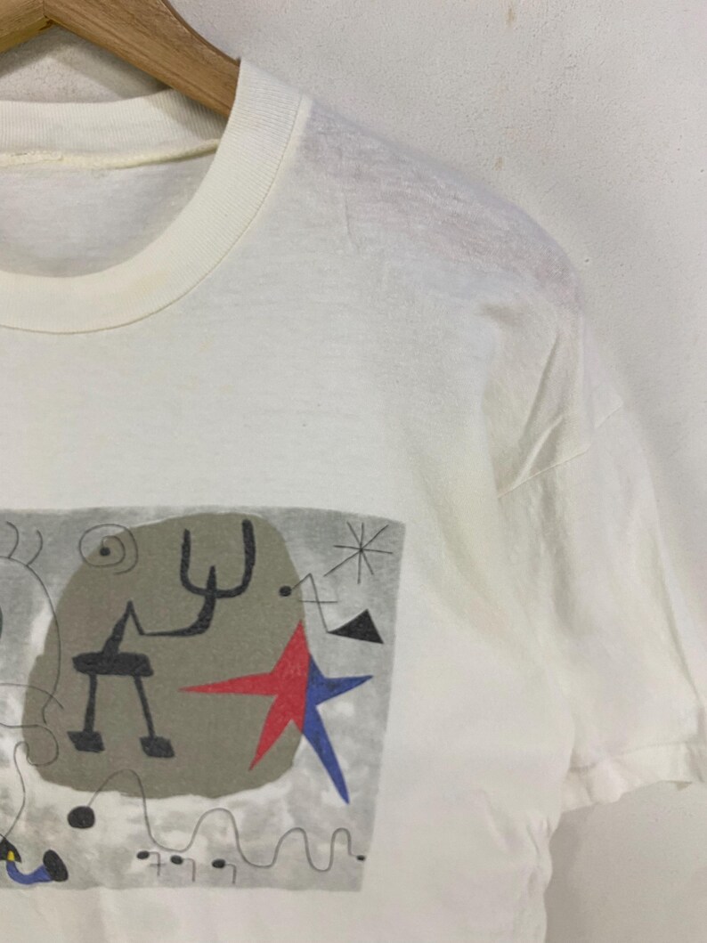 Puede incluir: Camiseta blanca con un dise&ntilde;o abstracto. El dise&ntilde;o incluye un fondo gris, formas negras, una estrella roja y una estrella azul. La camiseta est&aacute; hecha de un material suave y ligero.