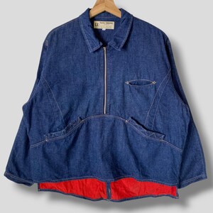 Op de afbeelding: Een blauw spijkerjasje met ritssluiting, twee voorzakken en een rode voering aan de onderkant. De jas hangt aan een houten hanger.
