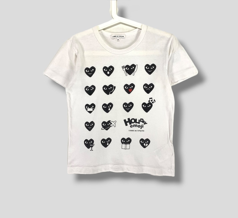 Puede incluir: Camiseta blanca con cuello redondo y mangas cortas. La camiseta presenta un patr&oacute;n de emojis negros en forma de coraz&oacute;n con diversas expresiones. El texto "HOLIDAY emoji" est&aacute; impreso debajo de los corazones. La marca "COMME des GAR&Ccedil;ONS" tambi&eacute;n es visible.