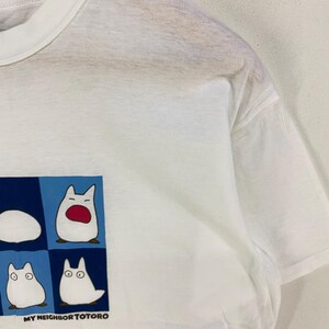 May include: White t-shirt featuring a graphic of four Totoro characters in a blue and white square. The text "MY NEIGHBOR TOTORO" is printed below the graphic. The shirt has short sleeves and a crew neck.
