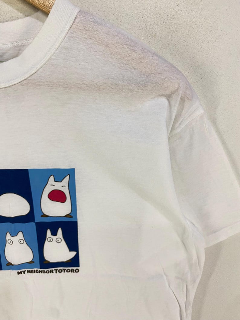 May include: White t-shirt featuring a graphic of four Totoro characters in a blue and white square. The text "MY NEIGHBOR TOTORO" is printed below the graphic. The shirt has short sleeves and a crew neck.