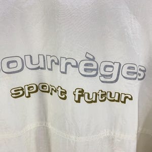 K&ouml;nnte beinhalten: Nahaufnahme einer wei&szlig;en Jacke mit dem Aufdruck "courr&egrave;ges sport futur" in Grau und Olivgr&uuml;n. Die Jacke hat eine leicht zerknitterte Textur.