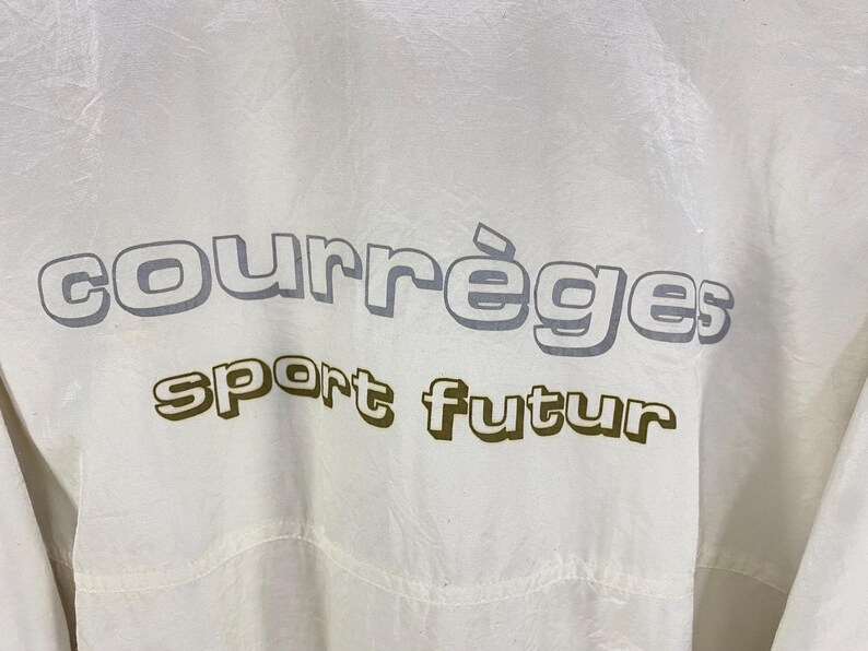 K&ouml;nnte beinhalten: Nahaufnahme einer wei&szlig;en Jacke mit dem Aufdruck "courr&egrave;ges sport futur" in Grau und Olivgr&uuml;n. Die Jacke hat eine leicht zerknitterte Textur.