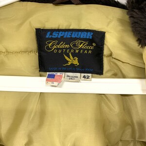 Puede incluir: Primer plano de la etiqueta de una chaqueta de abrigo L. Spiewak Golden Fleece de color beige. La etiqueta es negra con letras doradas y un gr&aacute;fico de p&aacute;jaro dorado. La etiqueta dice "MADE IN THE U.S.A. Since 1904". La chaqueta es de talla 42.