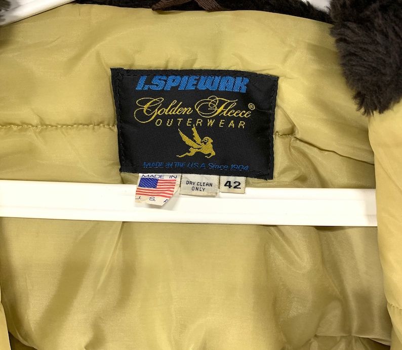 Puede incluir: Primer plano de la etiqueta de una chaqueta de abrigo L. Spiewak Golden Fleece de color beige. La etiqueta es negra con letras doradas y un gr&aacute;fico de p&aacute;jaro dorado. La etiqueta dice "MADE IN THE U.S.A. Since 1904". La chaqueta es de talla 42.