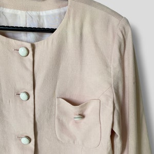 May include: A pale pink blazer with long sleeves and a single pocket on the left side. The blazer has a round neckline and is fastened with white buttons.