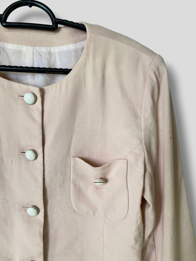 May include: A pale pink blazer with long sleeves and a single pocket on the left side. The blazer has a round neckline and is fastened with white buttons.