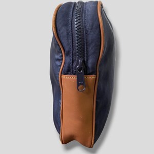 May include: A navy blue zippered pouch with brown leather accents. The pouch has a rectangular shape with a brown leather panel on one side and brown trim around the edges. The zipper is black.