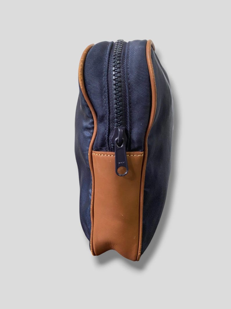 May include: A navy blue zippered pouch with brown leather accents. The pouch has a rectangular shape with a brown leather panel on one side and brown trim around the edges. The zipper is black.