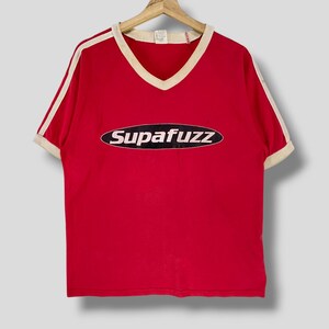 May include: Red V-neck t-shirt with white trim and stripes on the sleeves. The shirt features the text "Supafuzz" in a white, oval-shaped design with a navy blue outline. The shirt is hanging on a wooden hanger.