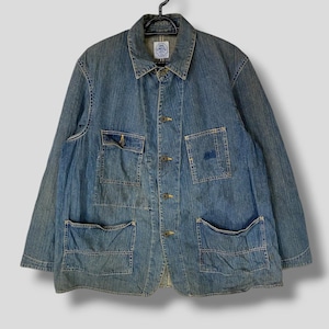 Puede incluir: Chaqueta vaquera azul claro con múltiples bolsillos delanteros y botones dorados. La chaqueta, colgada de una percha negra, tiene un cuello clásico. La tela muestra signos de desgaste, lo que aumenta su atractivo vintage. La chaqueta mide unos 76 cm de largo.