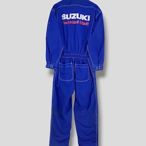Op de afbeelding: Blauw overall met witte stiksels en het Suzuki-logo op de rug. De overall heeft lange mouwen, een knoopsluiting en zakken op de borst en rug. De tekst "Technical Staff" staat onder het Suzuki-logo.