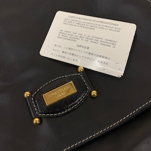 May include: Black leather handbag with a gold Courr&egrave;ges Paris emblem and gold-toned studs. A white authenticity card is on top, with text in French and English. The bag features white stitching.