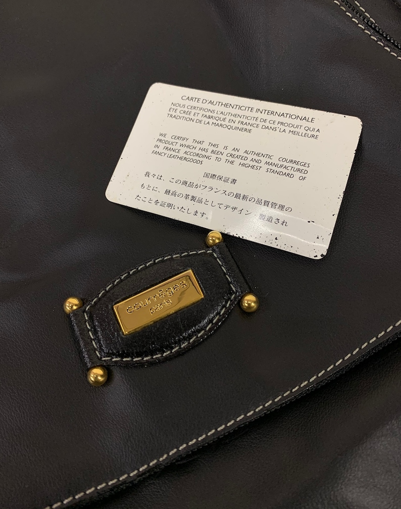 May include: Black leather handbag with a gold Courr&egrave;ges Paris emblem and gold-toned studs. A white authenticity card is on top, with text in French and English. The bag features white stitching.