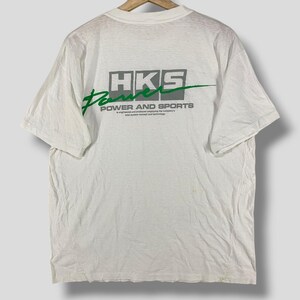 Puede incluir: Camiseta blanca con un logotipo HKS Power and Sports verde y gris en la parte posterior. El logotipo incluye el texto "HKS Power and Sports" y un swoosh verde.