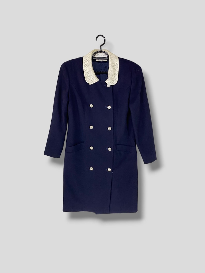 Puede incluir: Un vestido azul marino con cuello de encaje blanco y botones plateados. El vestido tiene cierre de doble botonadura y dos bolsillos.