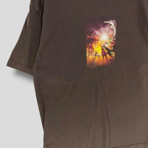 May include: A brown t-shirt with a vibrant graphic print. The print showcases a fantasy scene with figures, bright light, and a dynamic composition. The t-shirt has short sleeves and a crew neck.