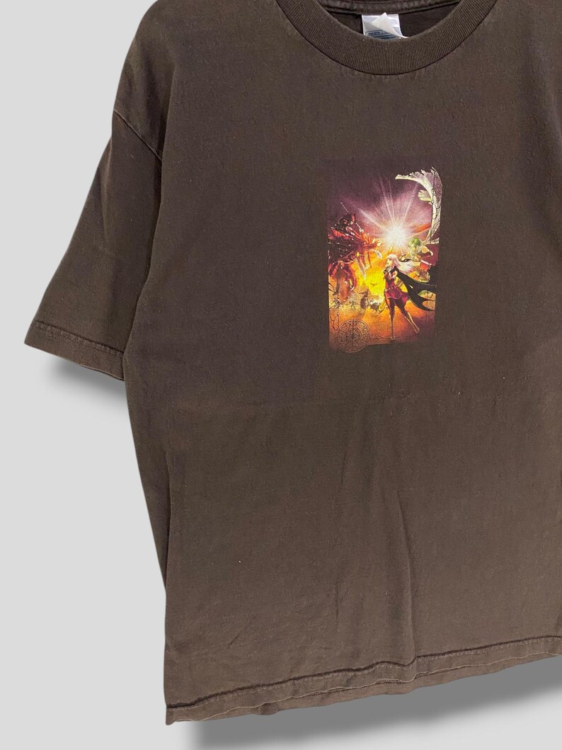 May include: A brown t-shirt with a vibrant graphic print. The print showcases a fantasy scene with figures, bright light, and a dynamic composition. The t-shirt has short sleeves and a crew neck.