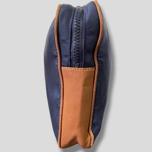 May include: A navy blue zippered pouch with brown leather accents. The pouch has a rectangular shape with a brown leather panel on the bottom. The zipper is dark blue, and the edges are trimmed with brown leather.