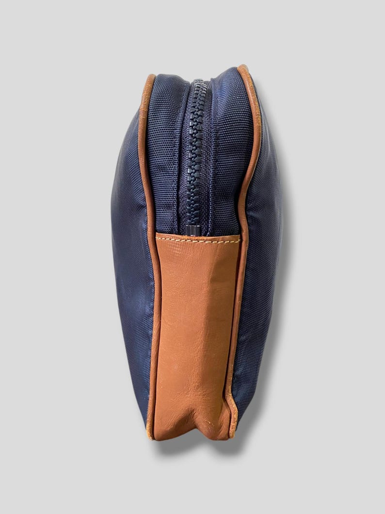 May include: A navy blue zippered pouch with brown leather accents. The pouch has a rectangular shape with a brown leather panel on the bottom. The zipper is dark blue, and the edges are trimmed with brown leather.