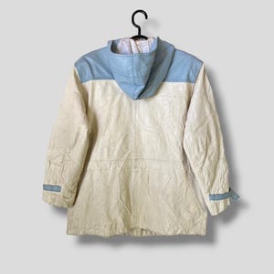 Puede incluir: Una chaqueta blanca con capucha y detalles de cuero azul en los hombros y las mangas. La chaqueta tiene una capucha con cord&oacute;n y una cremallera.