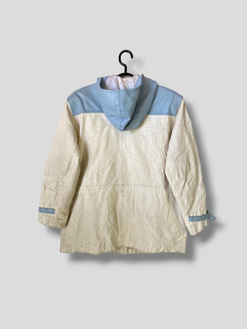 Puede incluir: Una chaqueta blanca con capucha y detalles de cuero azul en los hombros y las mangas. La chaqueta tiene una capucha con cord&oacute;n y una cremallera.